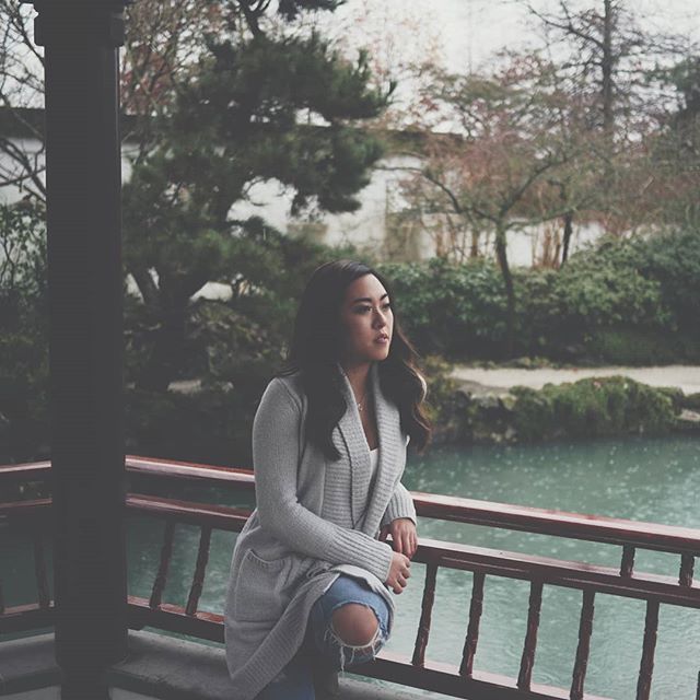 A woman in a cardigan and jeans gazes thoughtfully over a tranquil garden pond.
