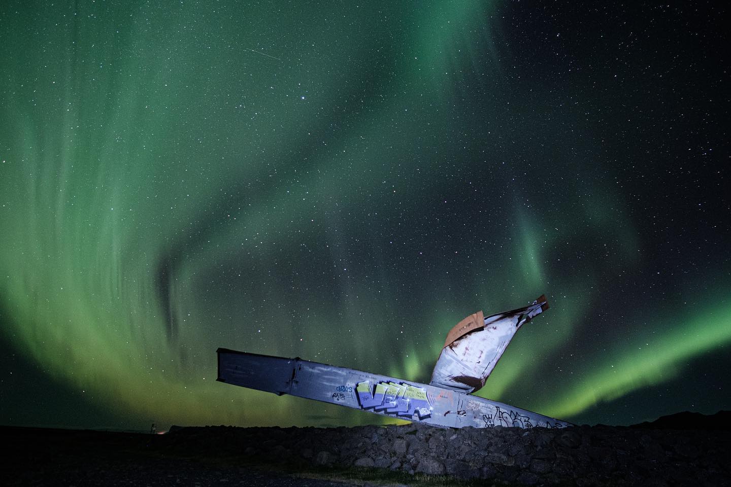 An airplane wreck sits under a stunning display of green Northern Lights in Iceland's night sky.