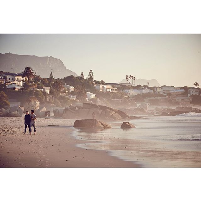A couple walks on a sandy beach with a town nestled along a coastline and mountain views.