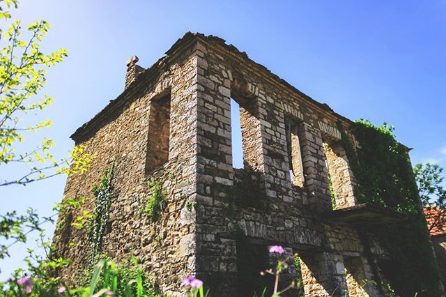 An old stone building stands in ruins, covered with greenery under a bright blue sky.