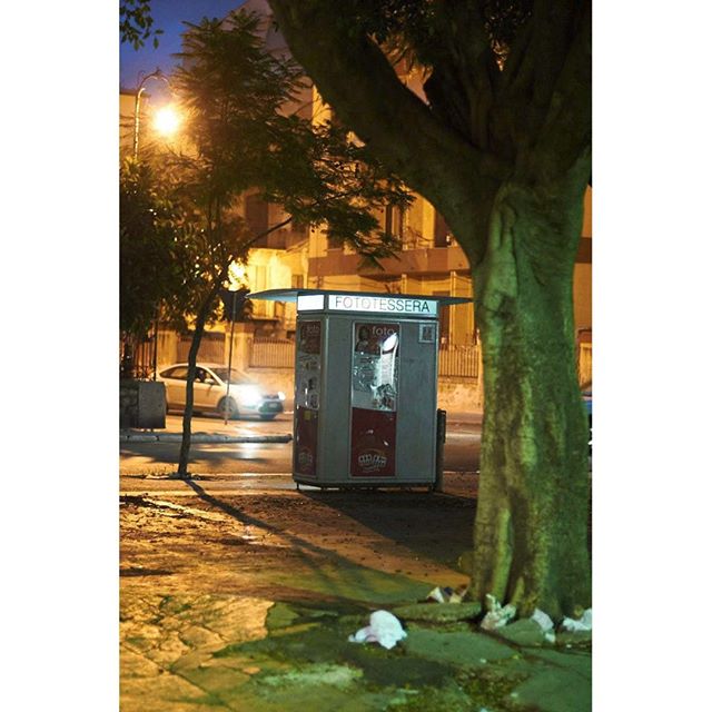 Night shot of a photobooth on a street, with a tree and car in the background.