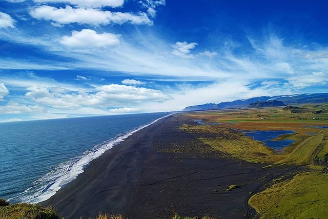 Panoramic view of a black sand beach meeting the ocean under a bright blue sky, capturing Iceland's serene coastal beauty.