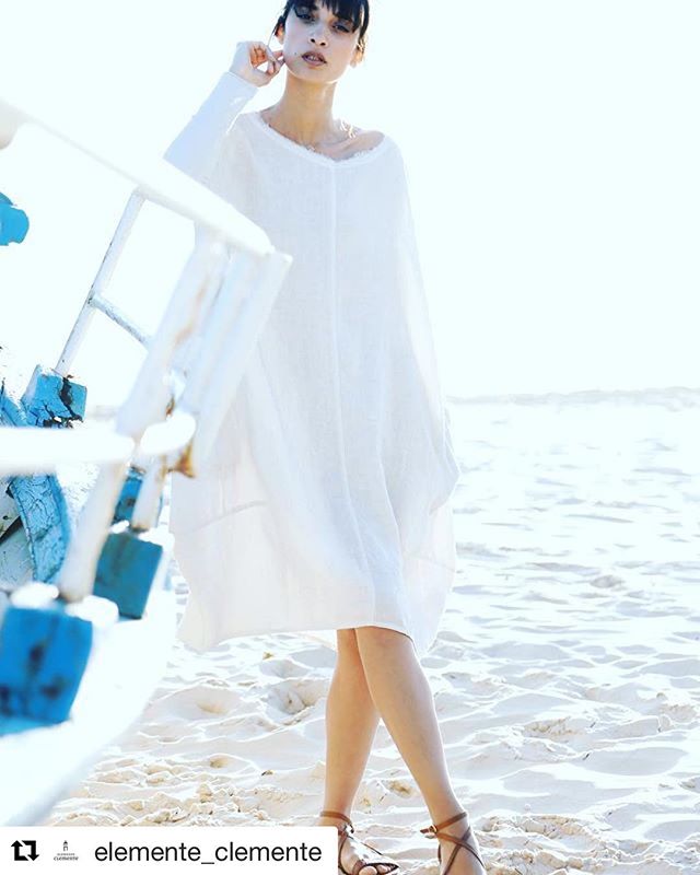 A woman in a white dress poses on a sandy beach by a blue and white boat.