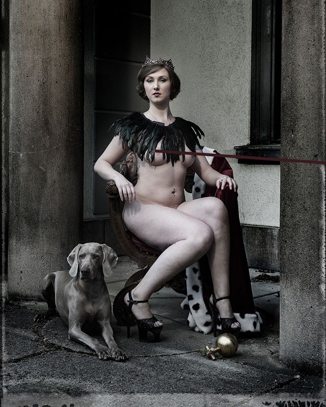 A nude woman with a crown and dog sits on a throne in an outdoor setting.