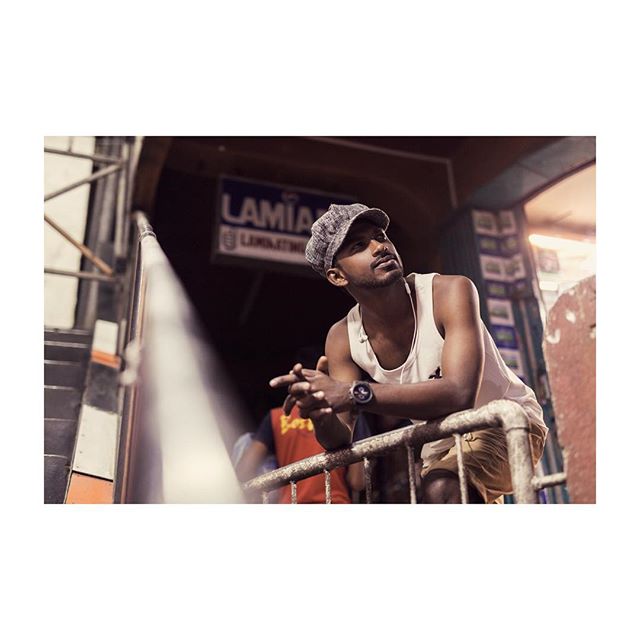 A man wearing a cap and tank top leans on a railing in an urban setting, looking thoughtful.