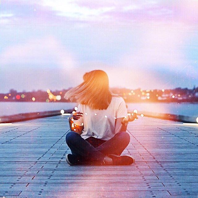 A woman meditates on a dock with dreamy lighting and soft focus, city lights glow in the background.