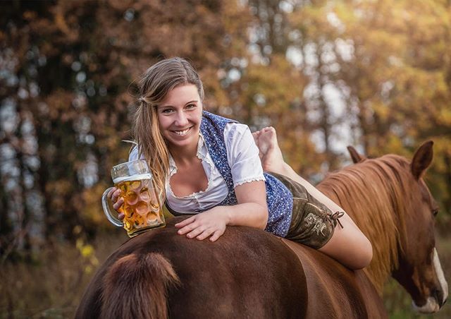 A smiling woman in traditional Bavarian dress lies on a horse holding a beer mug.