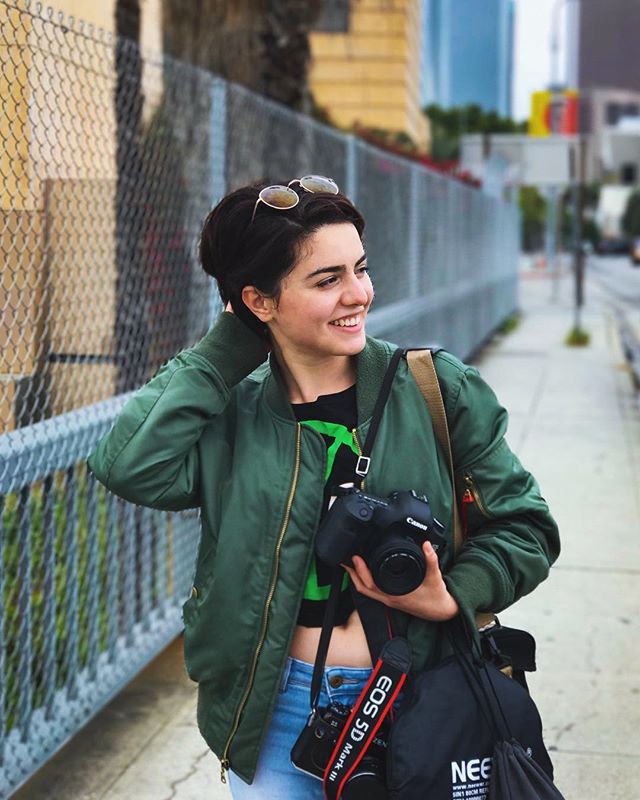 A smiling woman with a camera walks down a city street, showcasing an urban, travel lifestyle and street fashion.