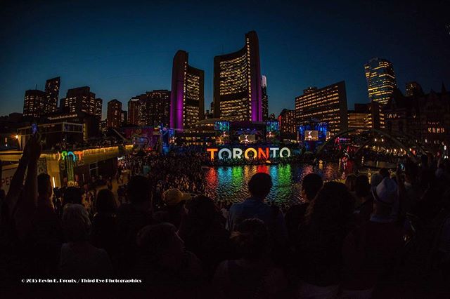 Night view of the Toronto skyline with a large crowd gathered for a concert event by a reflective lake.