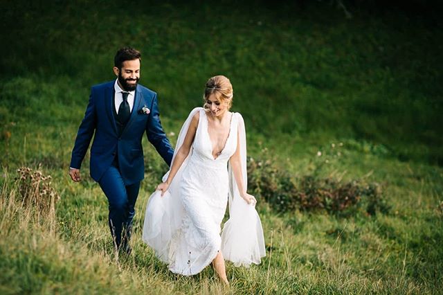 A smiling bride and groom walk hand-in-hand through a grassy field on their wedding day.