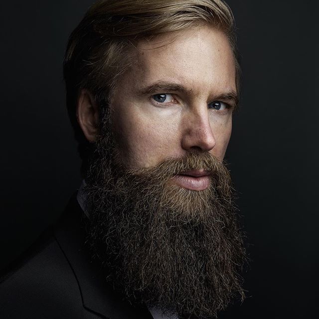 A man with a long beard poses in a professional portrait on a dark background.