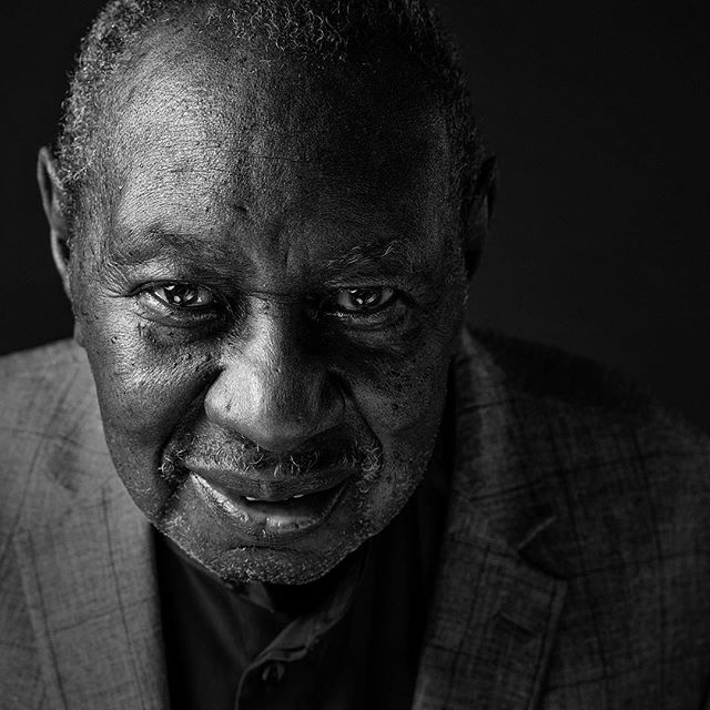 A black and white portrait of an elderly man in a suit, captured in a close-up shot with soft, dramatic lighting.