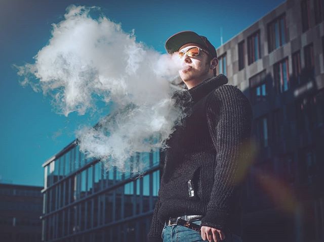 A man exhales a large cloud of vapor outdoors in front of a modern building while wearing sunglasses and a cap.