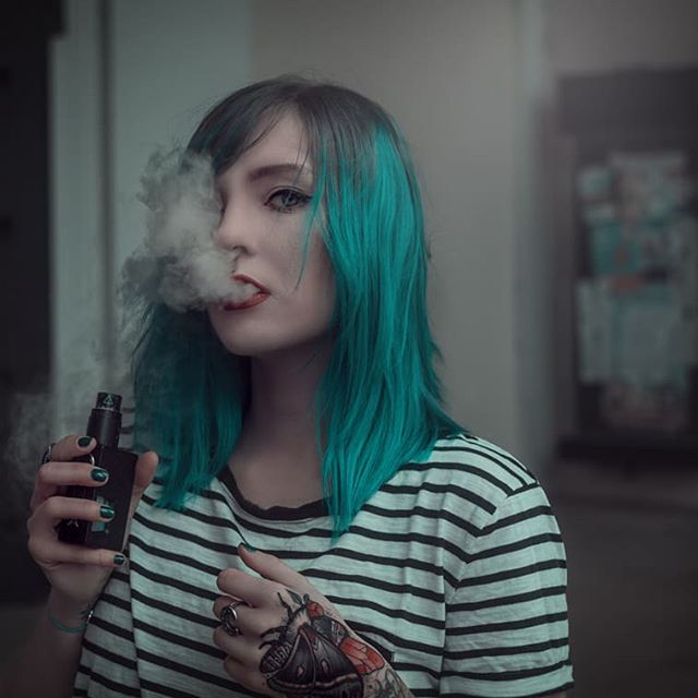 A woman with blue hair is vaping, blowing smoke out of her mouth outdoors.
