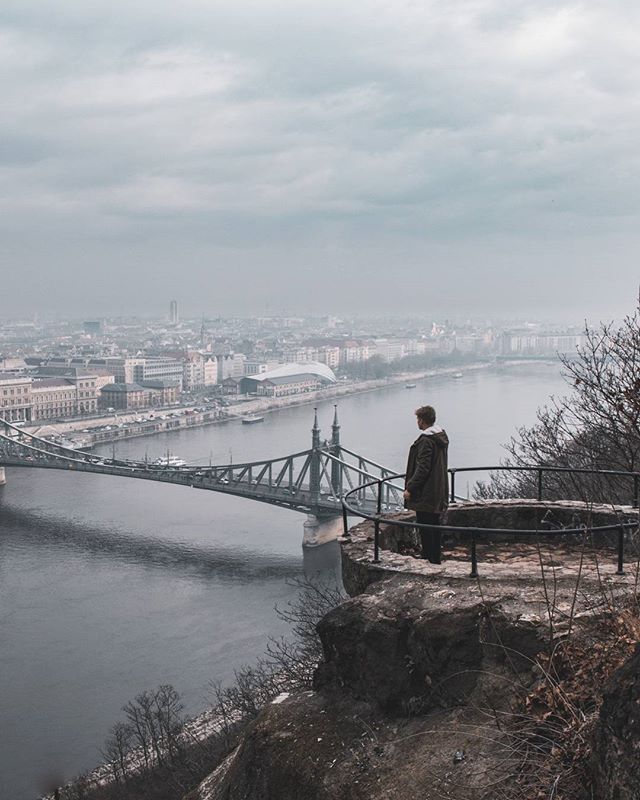A man stands on a cliff overlooking a bridge and cityscape on a cloudy day.