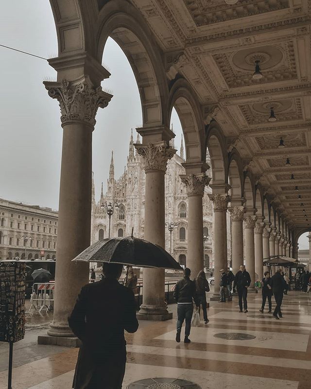 People stroll under an arcade with the Milan Cathedral visible in the background on a cloudy day.