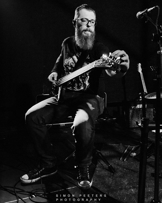 A bearded musician tunes his bass guitar on stage in a black and white concert photo.