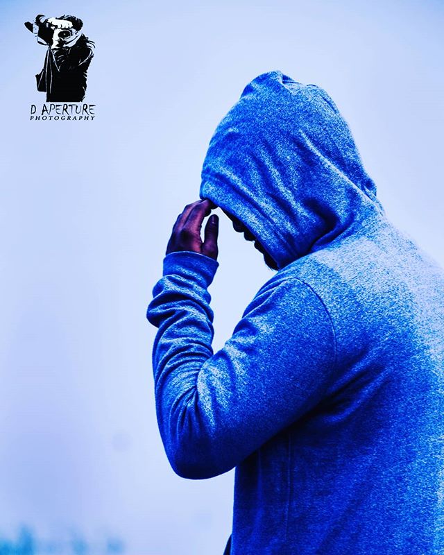 A person in a blue hoodie stands in a contemplative pose against a light blue sky.