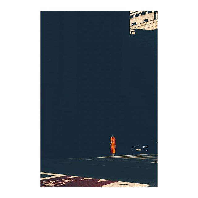 A lone woman in orange crosses a city street, a study in urban solitude and contrasting colors.