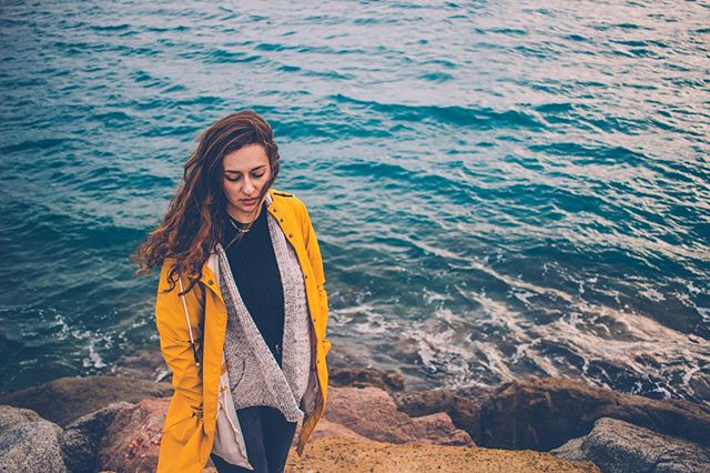 A woman in a yellow jacket stands near the ocean on a rocky coastline.