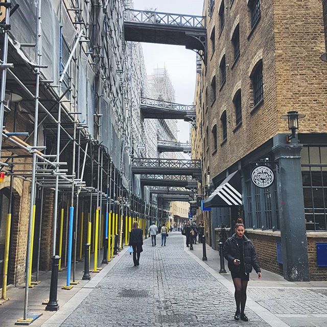 A woman walks down a narrow city alleyway lined with brick buildings and footbridges.