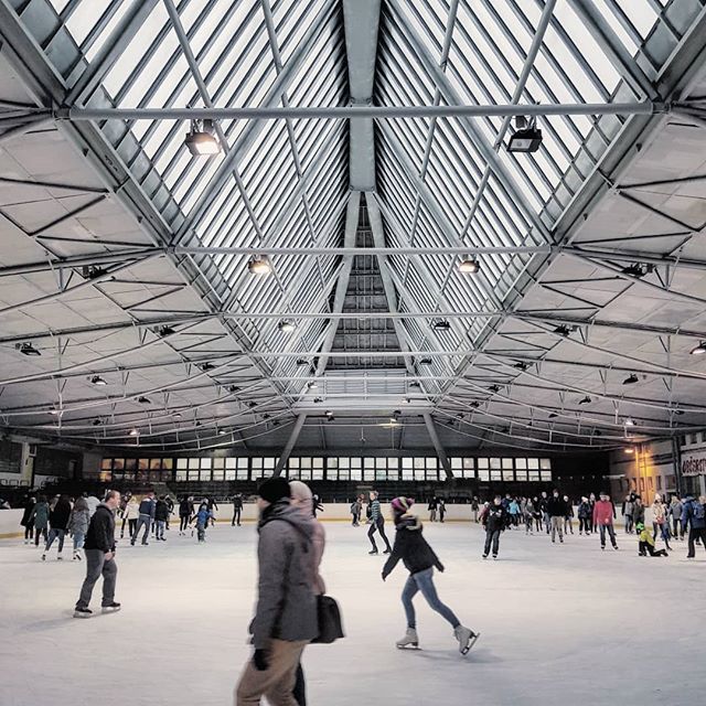 People skating on an indoor ice rink with a large metal roof and skylights.