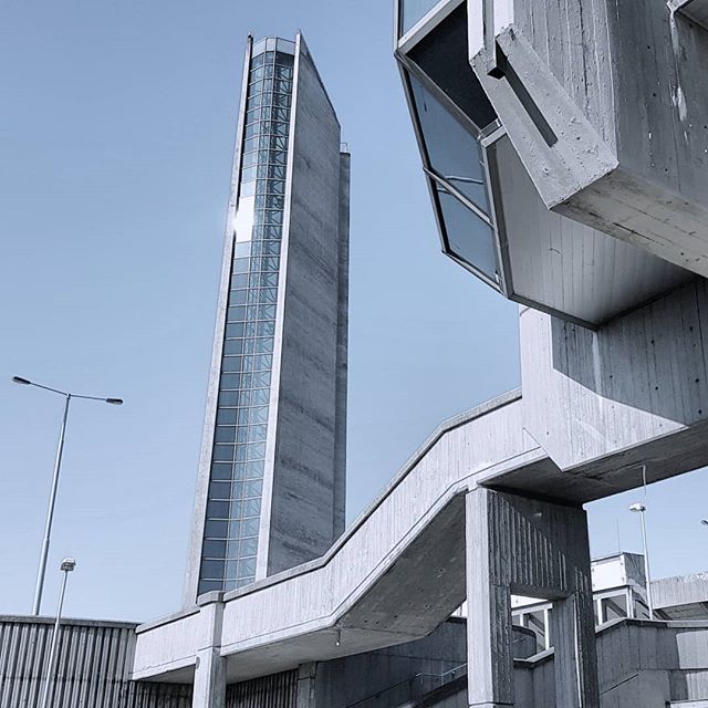 A concrete tower and connecting structure are set against a pale blue sky in a minimalist urban scene.