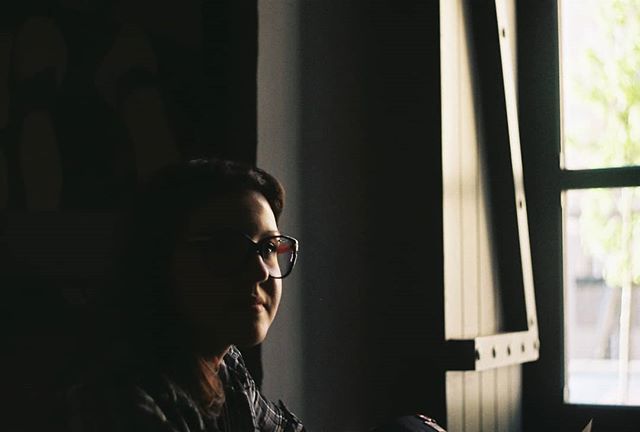 A woman wearing glasses gazes thoughtfully out the window in soft light.