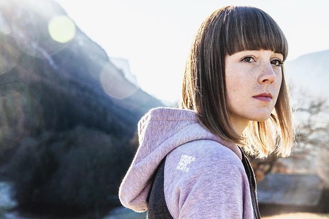 A brunette woman in a hoodie looks over her shoulder with a mountain backdrop.