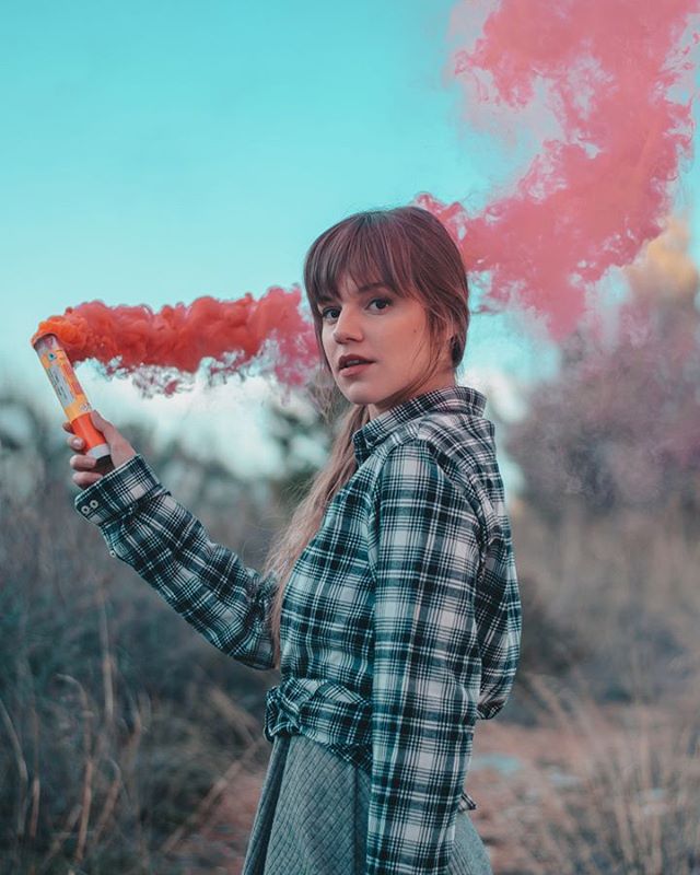 A young woman in a plaid shirt poses with a smoke grenade in a field.