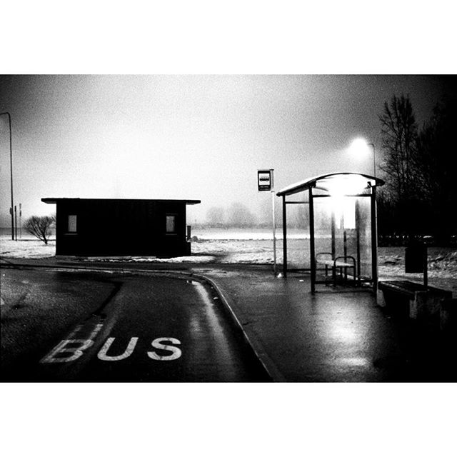 A black and white image of a bus stop shelter on a wet street at dusk.