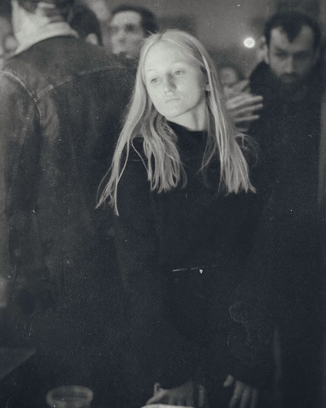 A vintage monochrome photo shows a woman in a crowd, at an indoor event, exuding a moody, emotional atmosphere.