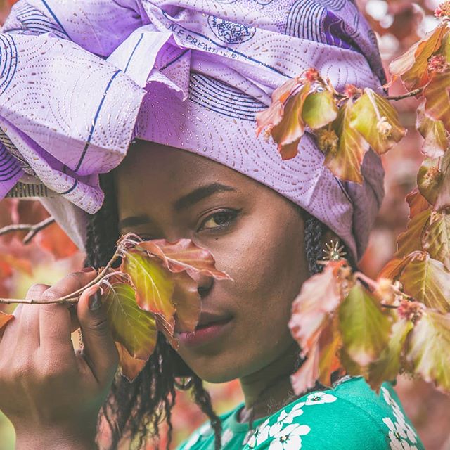 A woman wearing a purple headwrap poses with autumn leaves in a peaceful outdoor portrait.