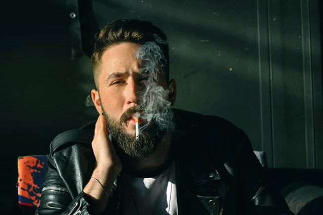 A bearded man in a leather jacket smokes a cigarette indoors in this moody editorial shot.