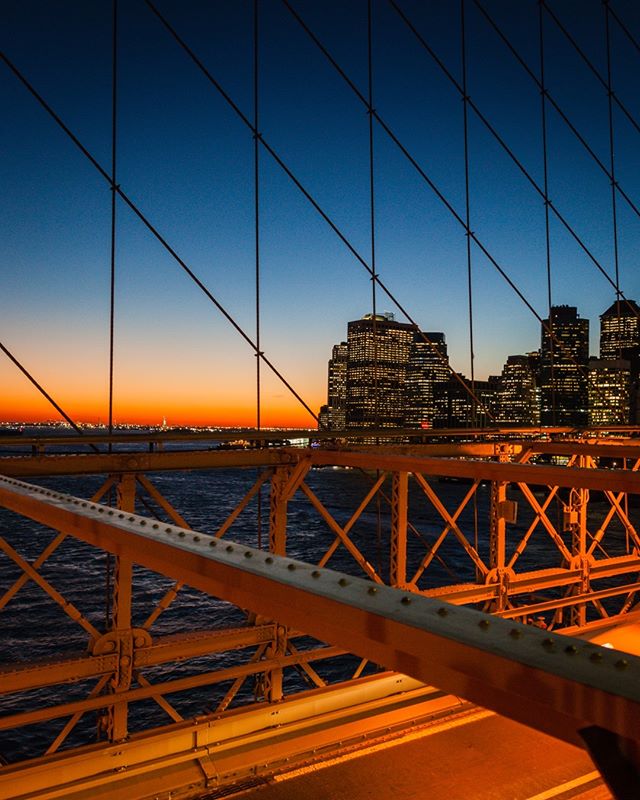 Illuminated buildings create a captivating cityscape view from the Brooklyn Bridge at night, with the East River reflecting the vibrant glow.