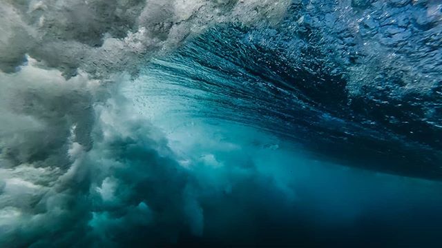 An ocean wave is viewed from underwater, with bright blue water and white foam.