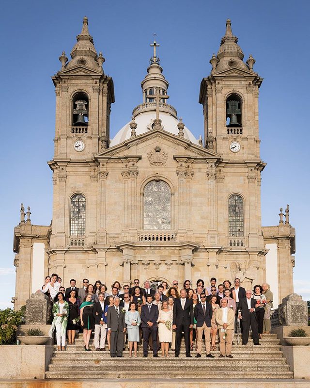 A large wedding group poses on church steps on a sunny day.