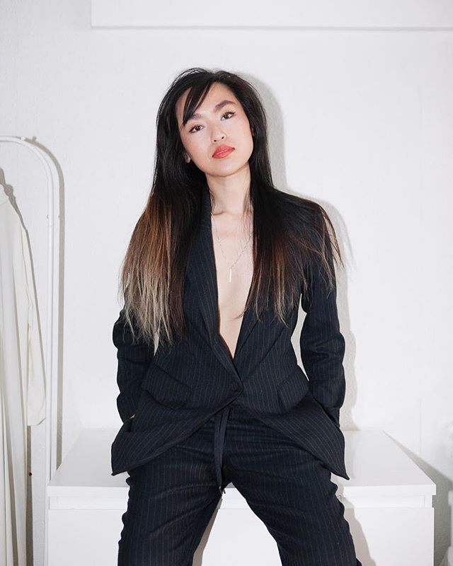 A brunette woman poses in a pinstripe suit and necklace, exuding confidence.