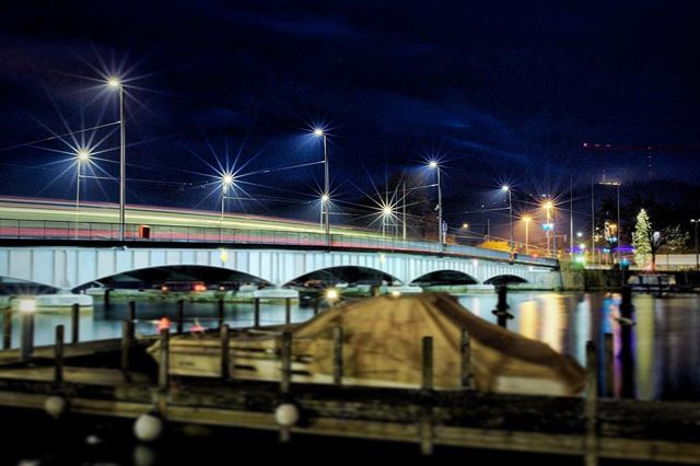 A bridge lit at night with long exposure light trails, reflects on still water in an urban cityscape.