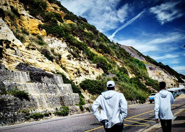 Two men walk along a road next to a tall cliff under a bright blue sky.