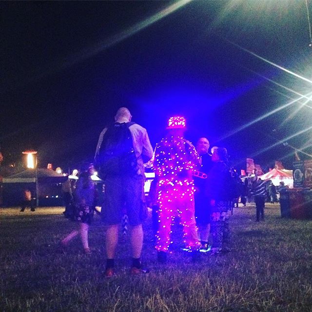 A group of people gather at night at a festival. One person stands out wearing a light-up LED suit.