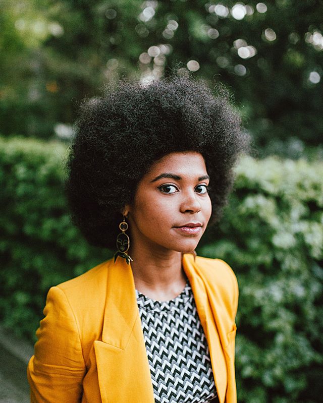 Stylish woman with afro and yellow blazer poses outdoors looking at the camera.