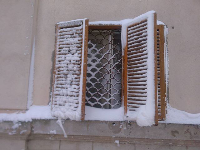 A window with wooden shutters is partially covered in snow, creating a serene winter scene.