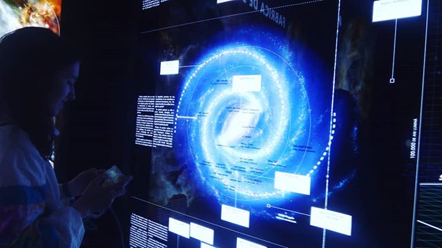 Woman looks at a digital display of a galaxy with text labels in a dark museum exhibit.