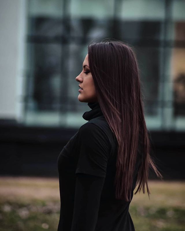 A woman with long brown hair poses in profile in an urban outdoor setting.