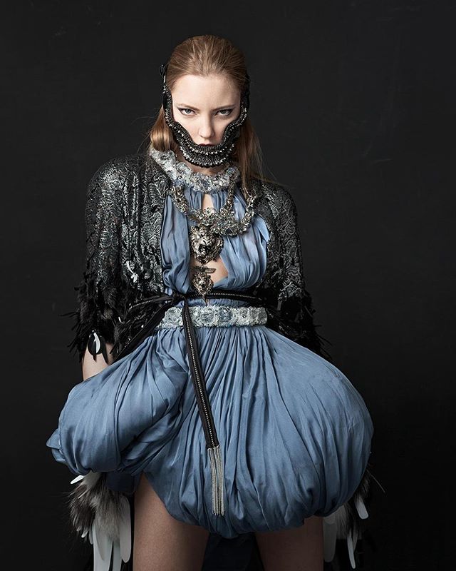 A fashion model poses in an avant-garde blue dress and a detailed face mask against a black background.