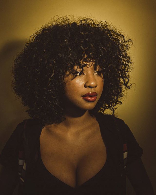 A stunning woman with curly hair and red lipstick poses in a studio with soft, warm lighting for a beauty campaign.
