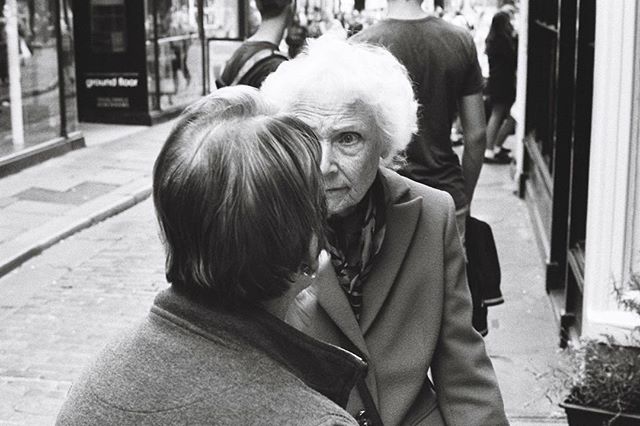 An elderly woman with white hair stares intently while standing on a city street in a black and white photograph.