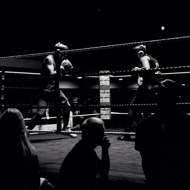Two boxers compete in a dimly lit boxing ring, surrounded by an audience in this vintage black and white image.