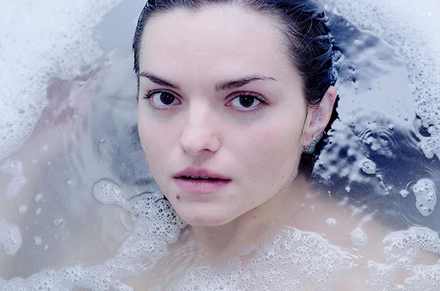 A woman is relaxing in a bubble bath looking directly at the viewer with a calm and relaxed expression.